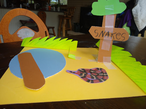 Child's craft project of a snake habitat with paper grass, a pond, a tree, and a sign saying 'SNAKES'.